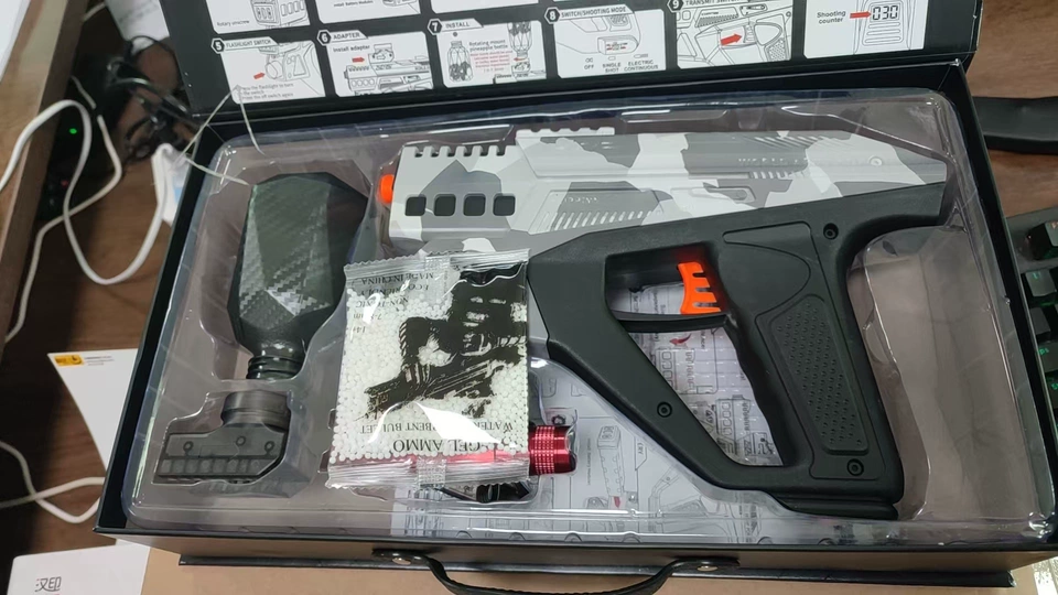 Boxed gel-blaster kit showing a gray camo pistol grip blaster, carbon-fiber-style hopper, bag of white gel beads, and red metal muzzle attachment neatly arranged in a clear plastic tray.